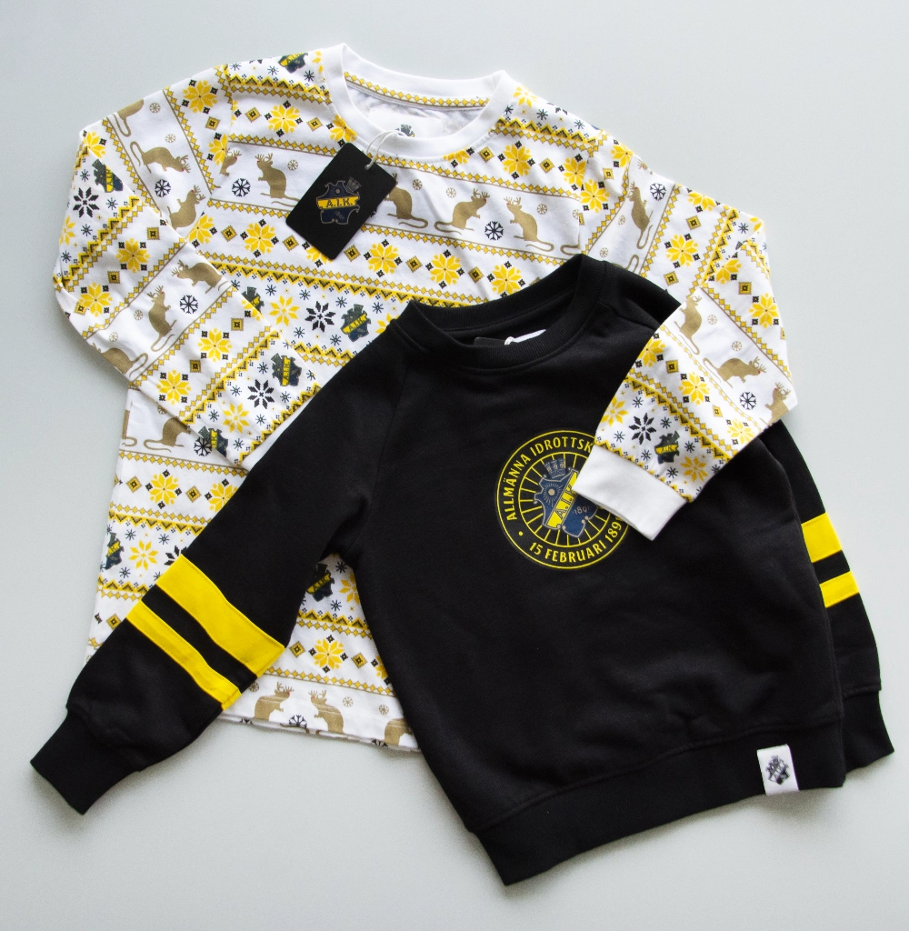  Två AIK barntröjor på ett ljust grått underalag: en vit med ren- och snöflingsmönster i gult och svart, och en svart med gula ärmränder och AIK-klubbmärket på bröstet